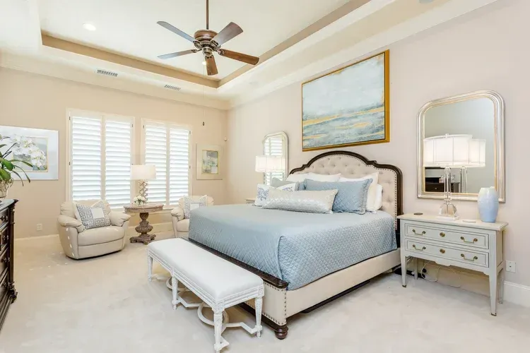 Bright bedroom with a blue bed, white bench, armchair, dresser, and ceiling fan