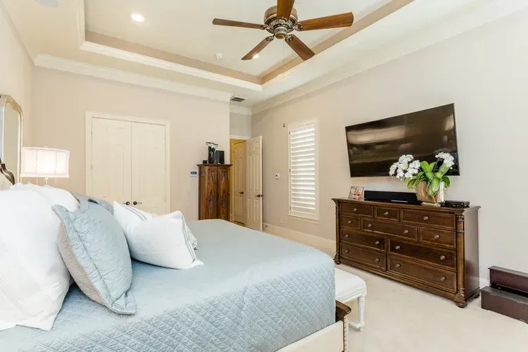 Bright bedroom with light blue bedspread, wall-mounted TV, dresser, ceiling fan, and beige walls