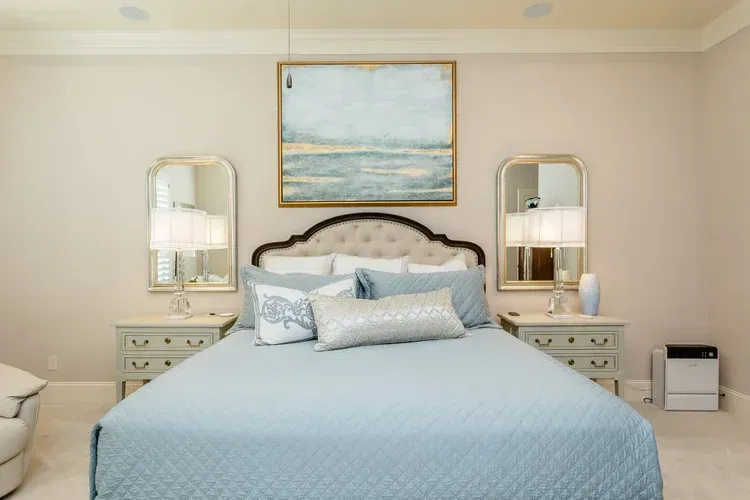 Elegant bedroom with a light blue bedspread, mirrored nightstands, lamps, and a seascape painting above the headboard
