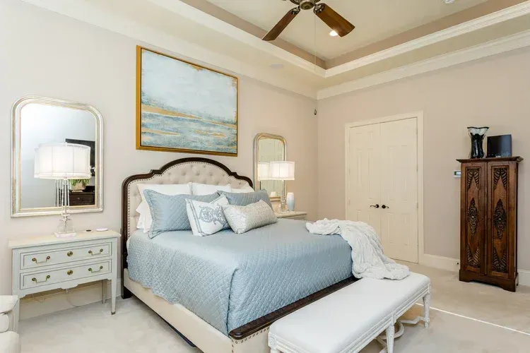 Bright bedroom with a light blue bedspread, white furniture, mirrors, and a framed seascape painting