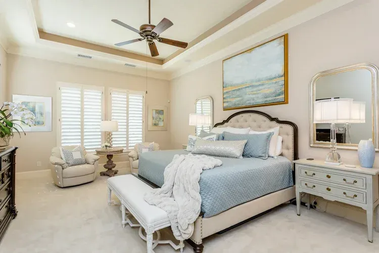Bright coastal-style bedroom with blue bed, white bench, and ceiling fan near shuttered windows