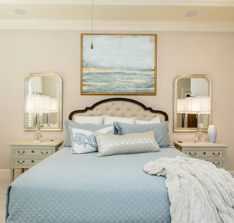 Elegant bedroom with a blue bed, white throw, mirrored nightstands, and a coastal painting above the headboard