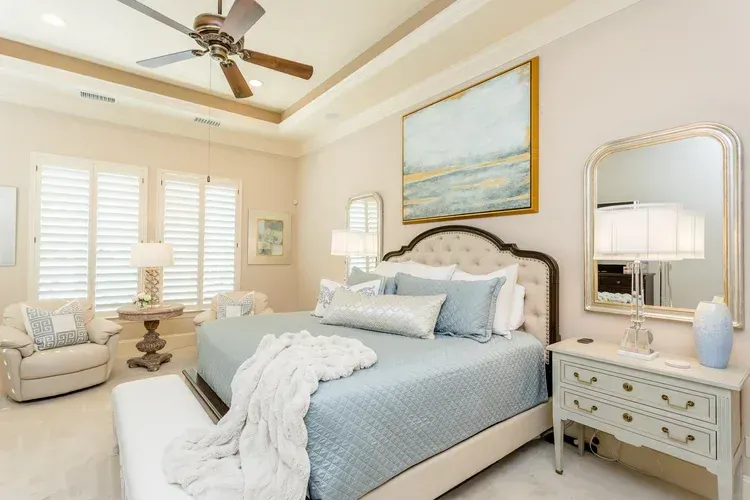 Bright cream bedroom with blue-accented bed, mirrored dresser, lamp, and ceiling fan