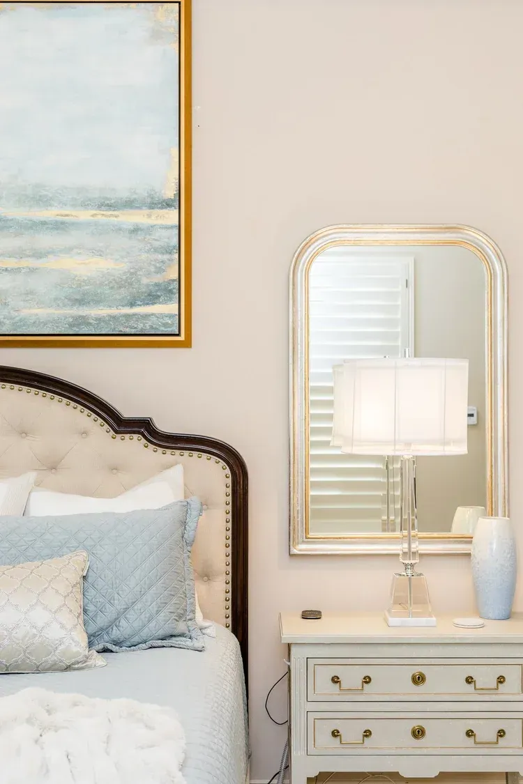 Elegant bedroom with framed art, a mirror, lamp, and white dresser beside a bed with light blue bedding