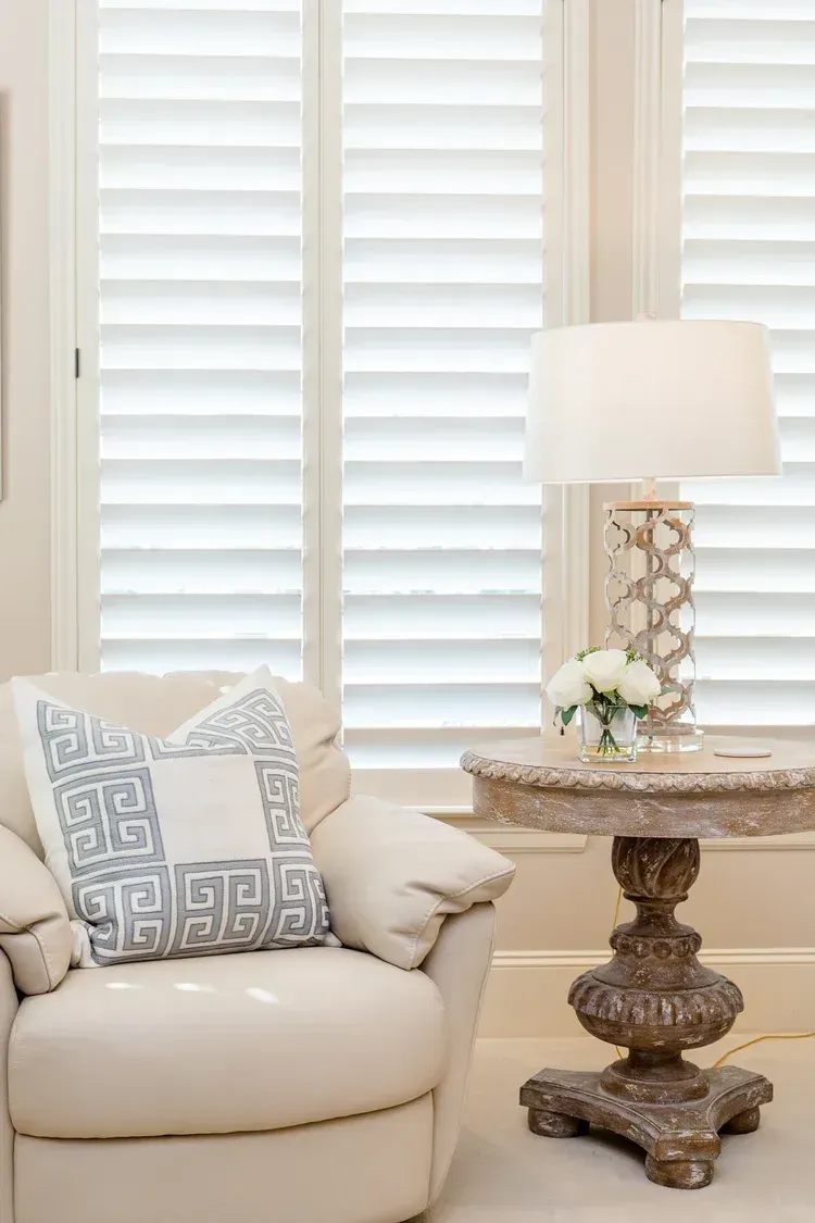 Cream armchair with patterned pillow beside ornate side table and lamp in a bright room with white shutters