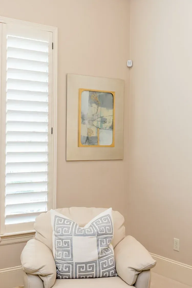 Cozy corner with cream armchair, patterned pillow, framed abstract art, and a window with white shutters