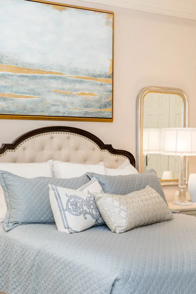 Elegant bedroom with a tufted bed, blue pillows, gold-framed art, and a mirrored bedside lamp.