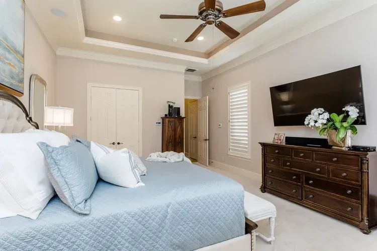 Bright bedroom with a light blue bedspread, dark wood dresser, wall-mounted TV, and ceiling fan.
