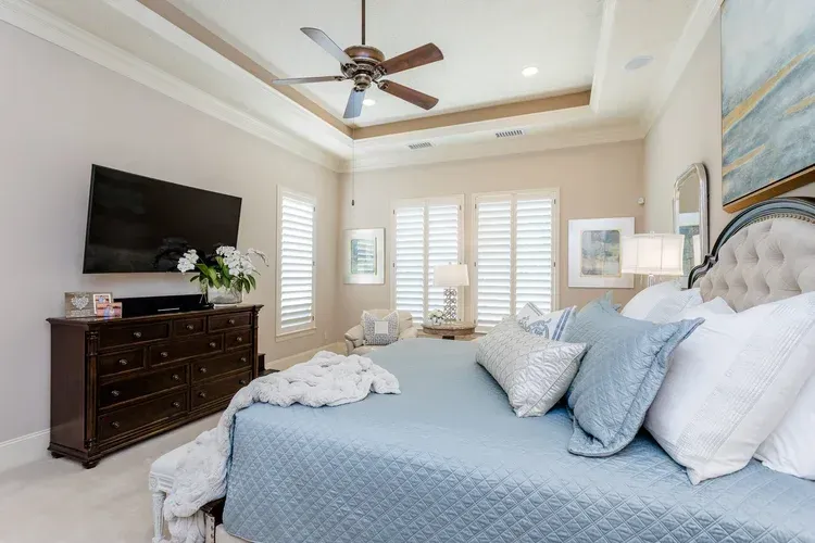 Bright bedroom with a large bed, wall-mounted TV, dark dresser, and windows with shutters and a ceiling fan.