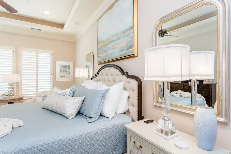 Bright bedroom with a tufted bed, pale blue bedding, wall art, mirror, and table lamp beside a window with shutters
