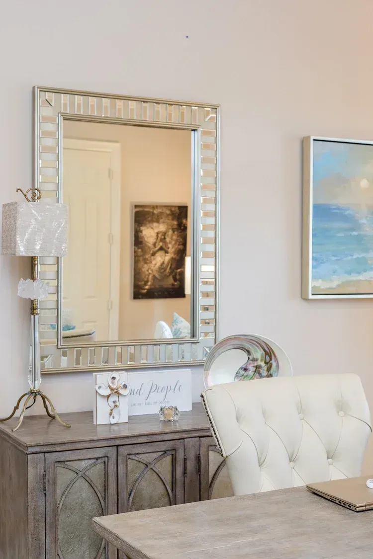 Decorative mirror above a console table, with a tufted white chair and framed wall art in a light room