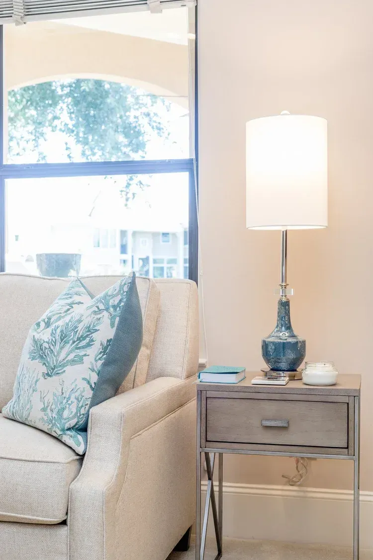 Cozy living room corner with cream armchair, blue patterned pillow, mirrored side table, and white lamp by a window