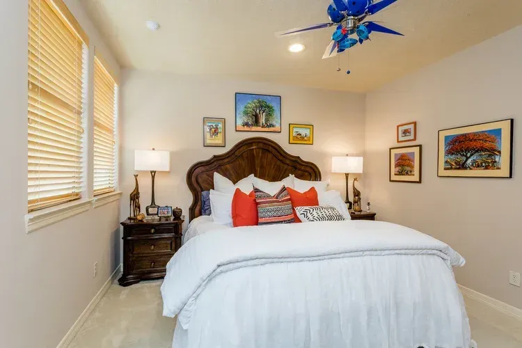 Bright bedroom with carved wooden bed, white bedding, red pillows, bedside lamp, and framed wall art