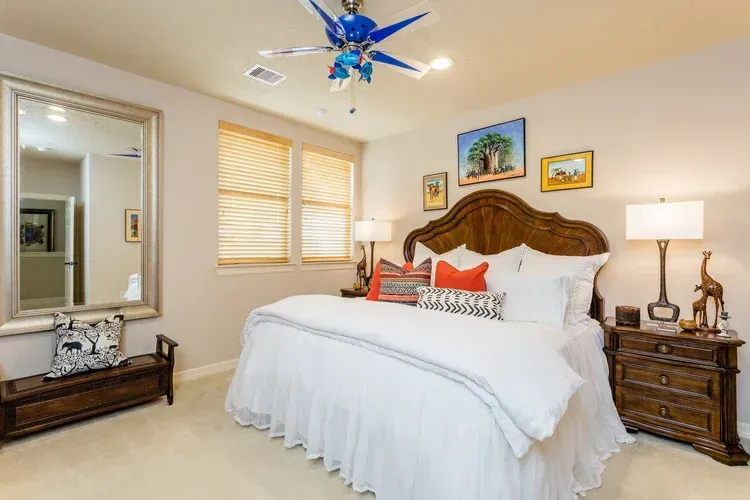 Bright bedroom with ornate bed, blue ceiling fan, bedside lamps, mirror, and framed wall art