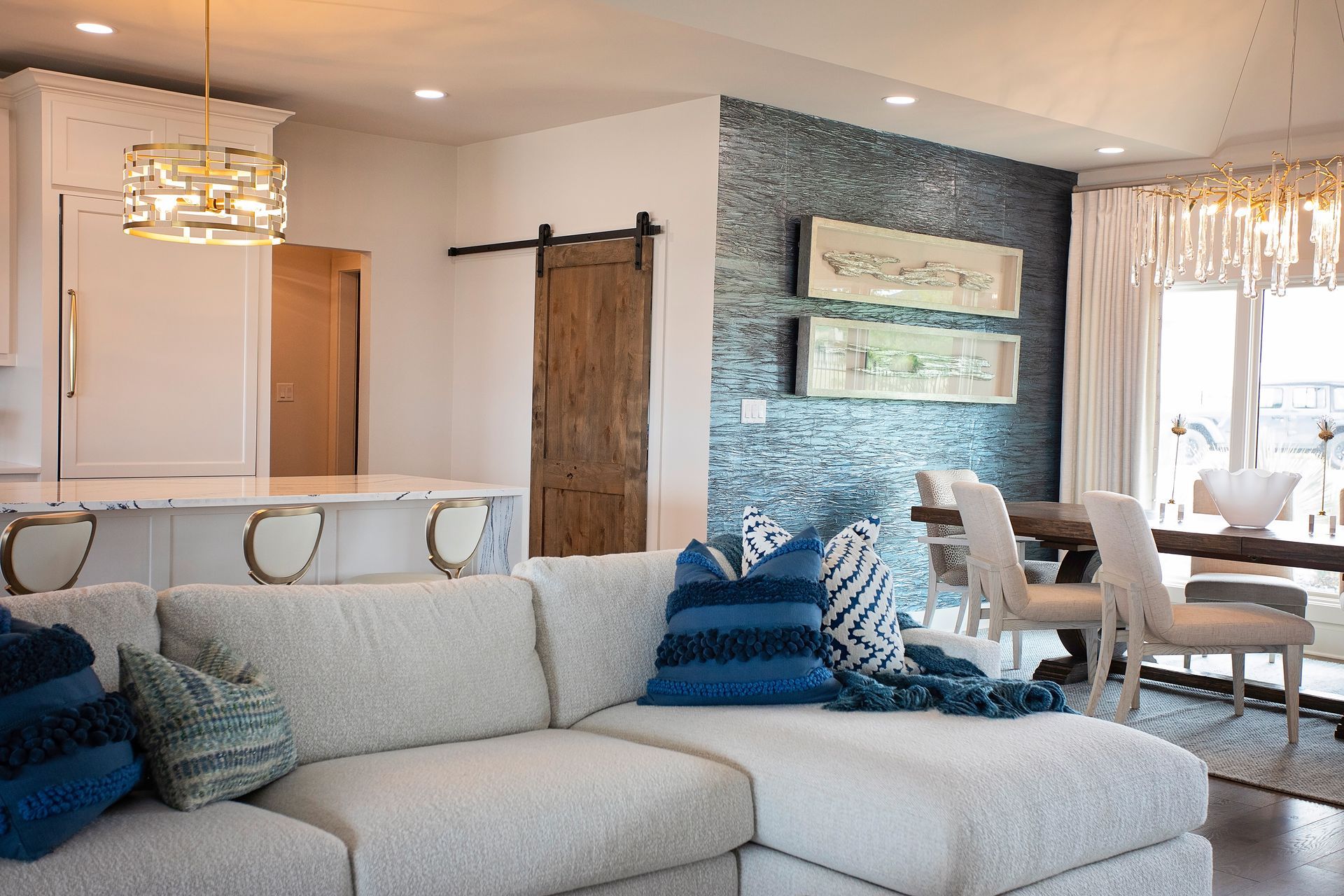 Bright open-concept living room and kitchen with gray sectional, blue pillows, island stools, and barn door