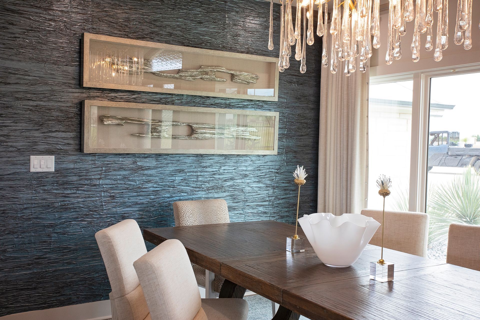 Modern dining room with dark stone wall, glass table, cream chairs, and chandelier near large windows