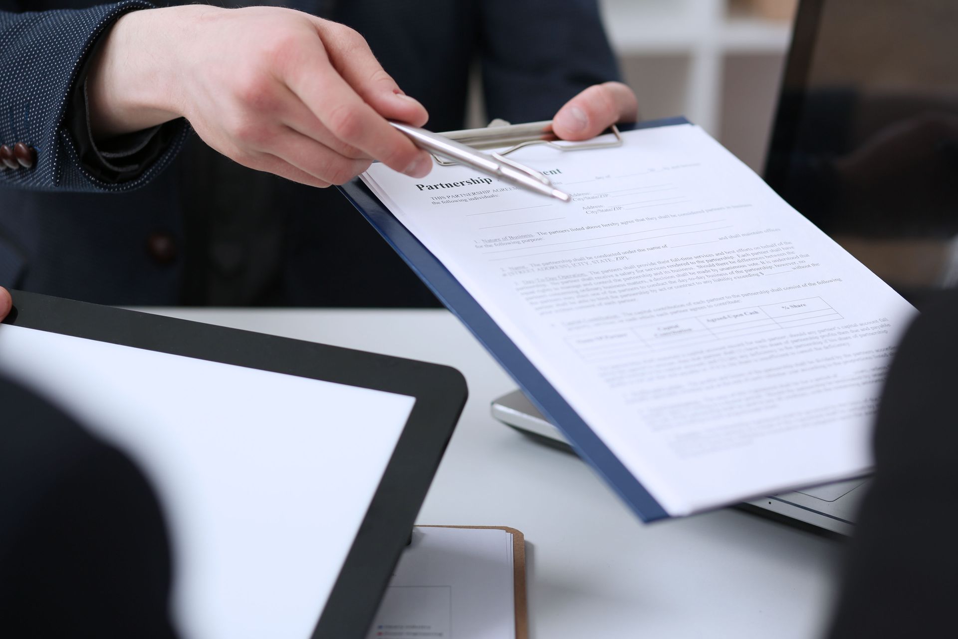 Man in suit points to document on a clipboard during a meeting, showing details to colleagues.