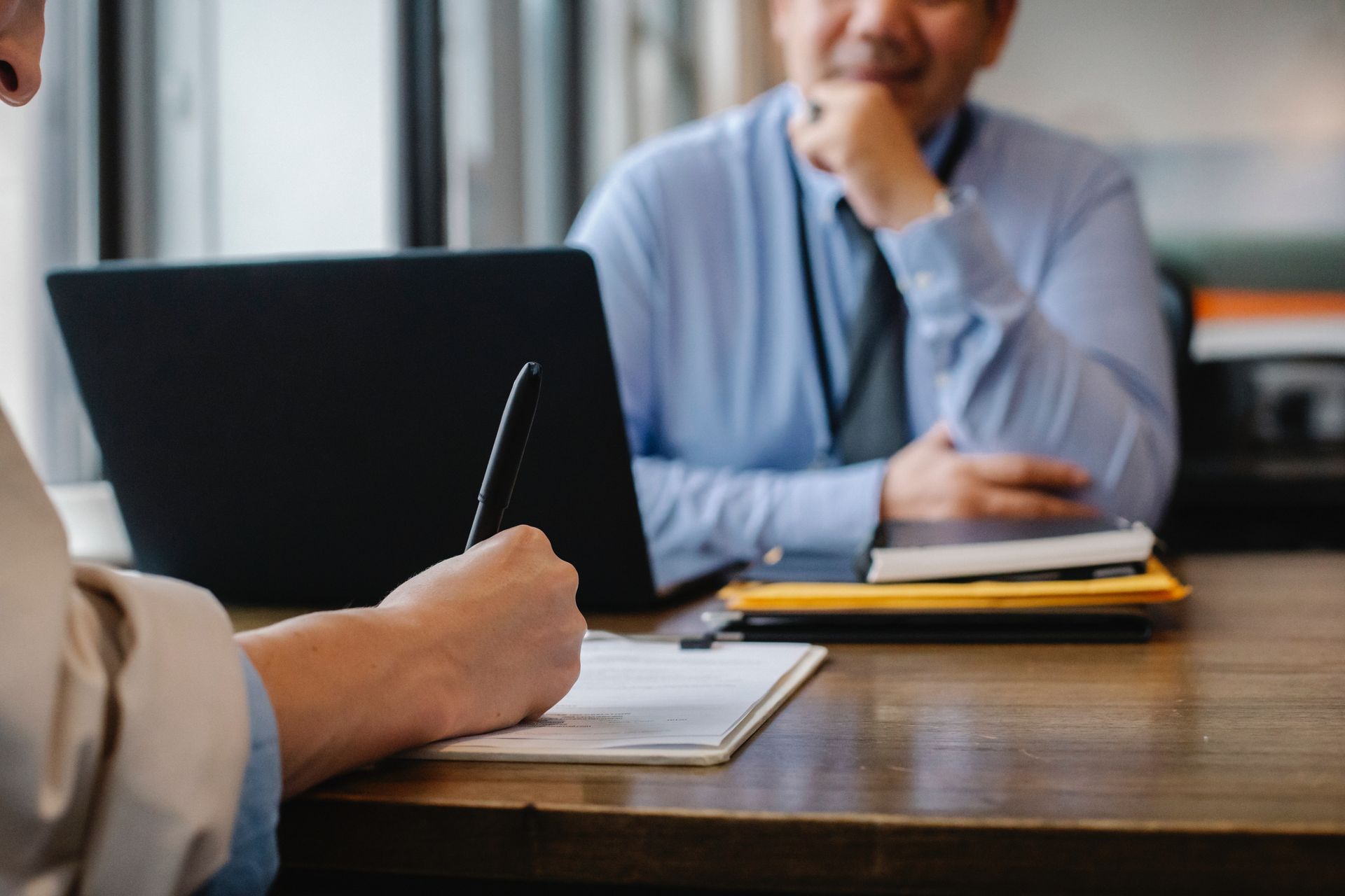 A person writes on a clipboard across from a person sitting at a table with a laptop, reflecting a professional meeting.