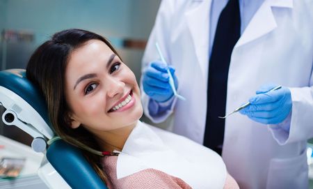 A woman is sitting in a dental chair smiling while a dentist examines her teeth.