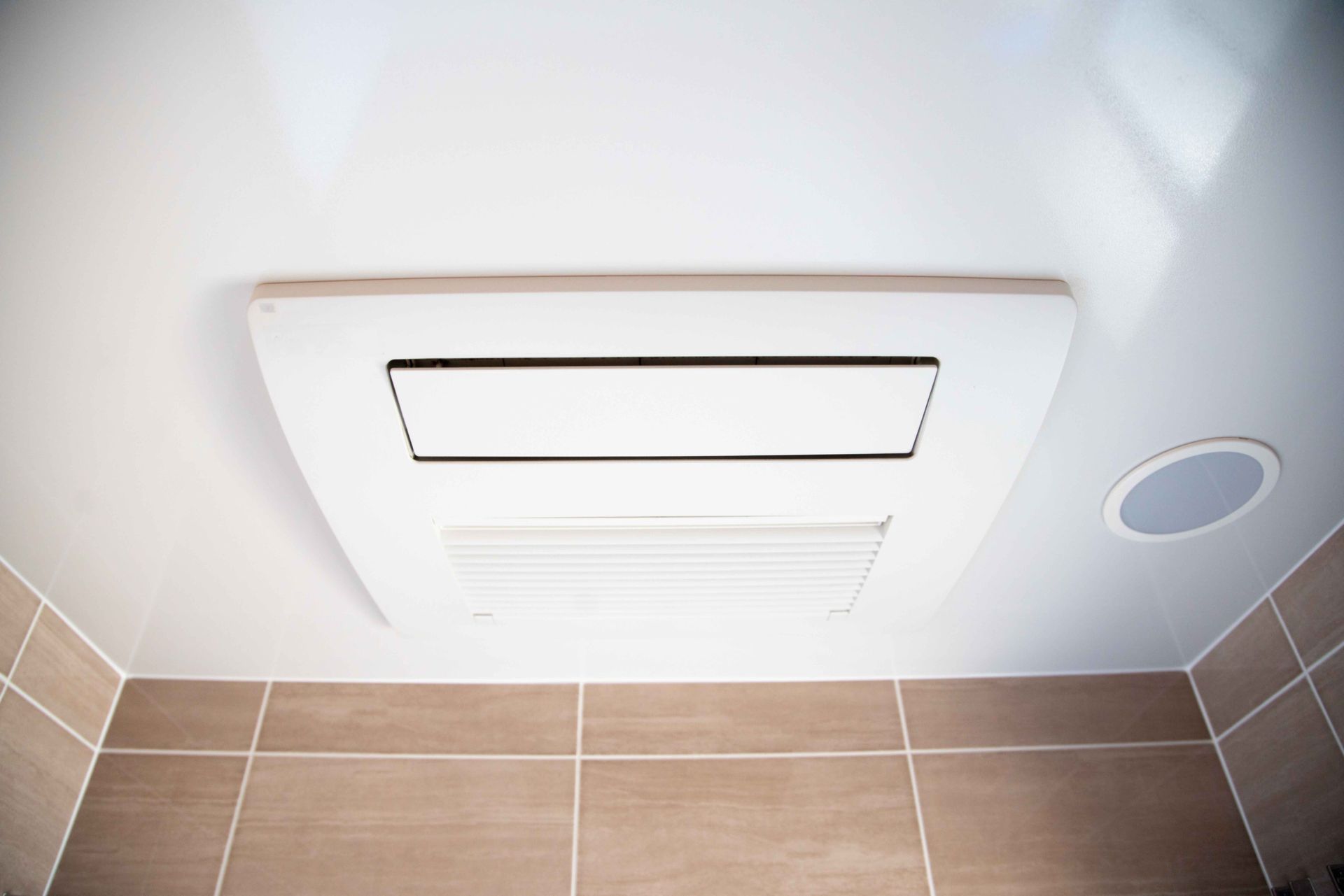 Bathroom ceiling vent and light fixture above tan tiled wall and floor.