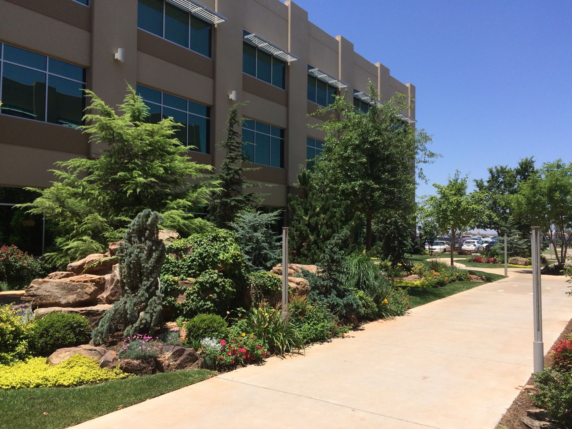 Building exterior with landscaped rock garden, trees, flowers, and a walkway on a sunny day.