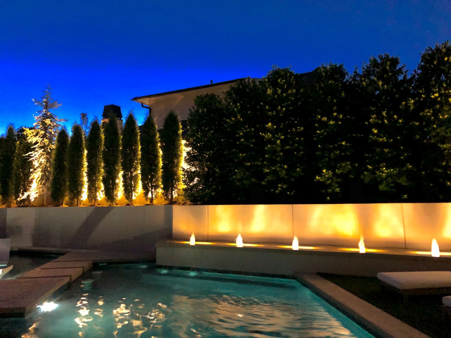 Pool area at night illuminated by lights, featuring a wall, trees, and a clear blue sky.