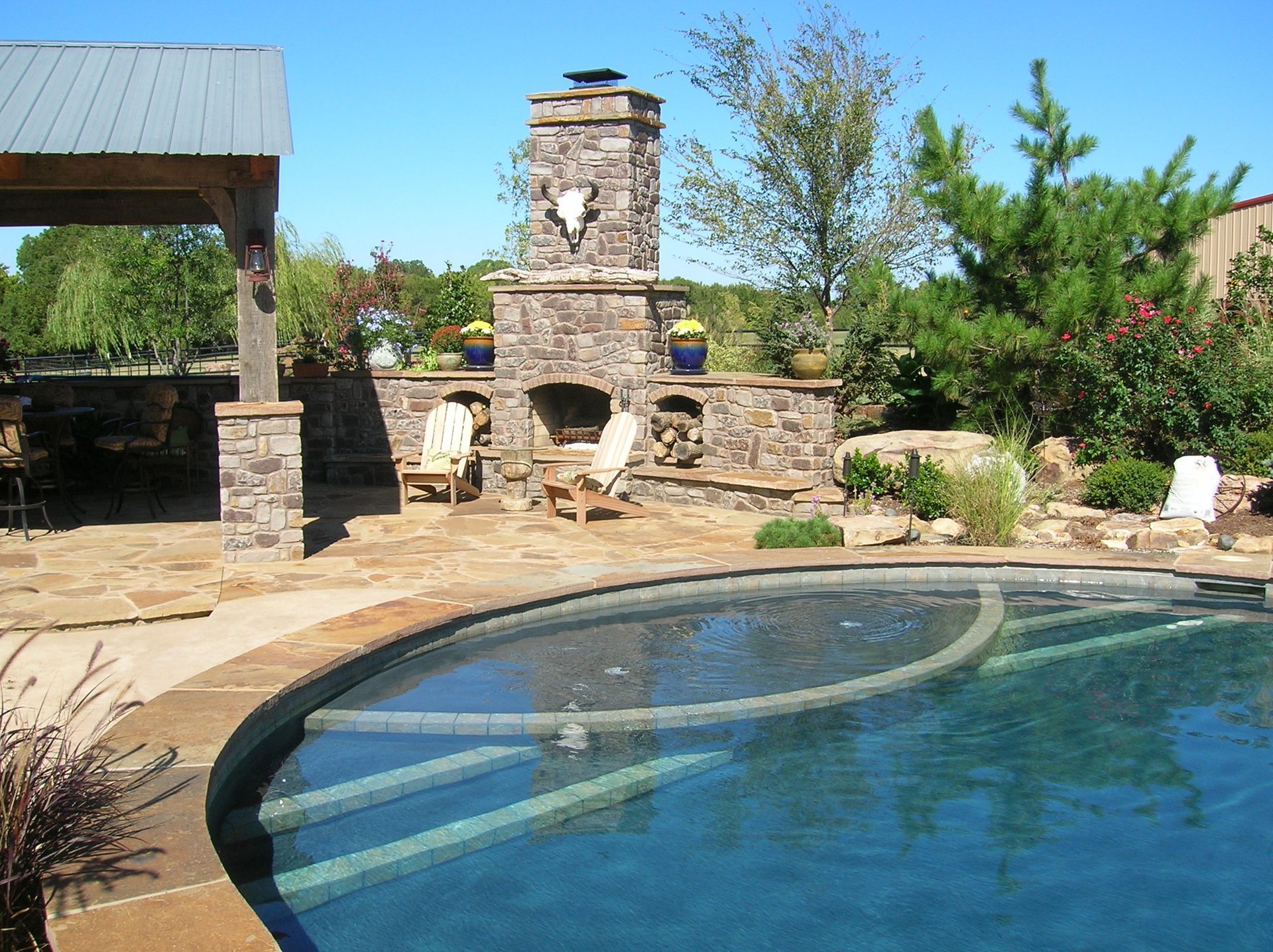 Stone fireplace and pool in a backyard setting, with wooden chairs and blue sky.