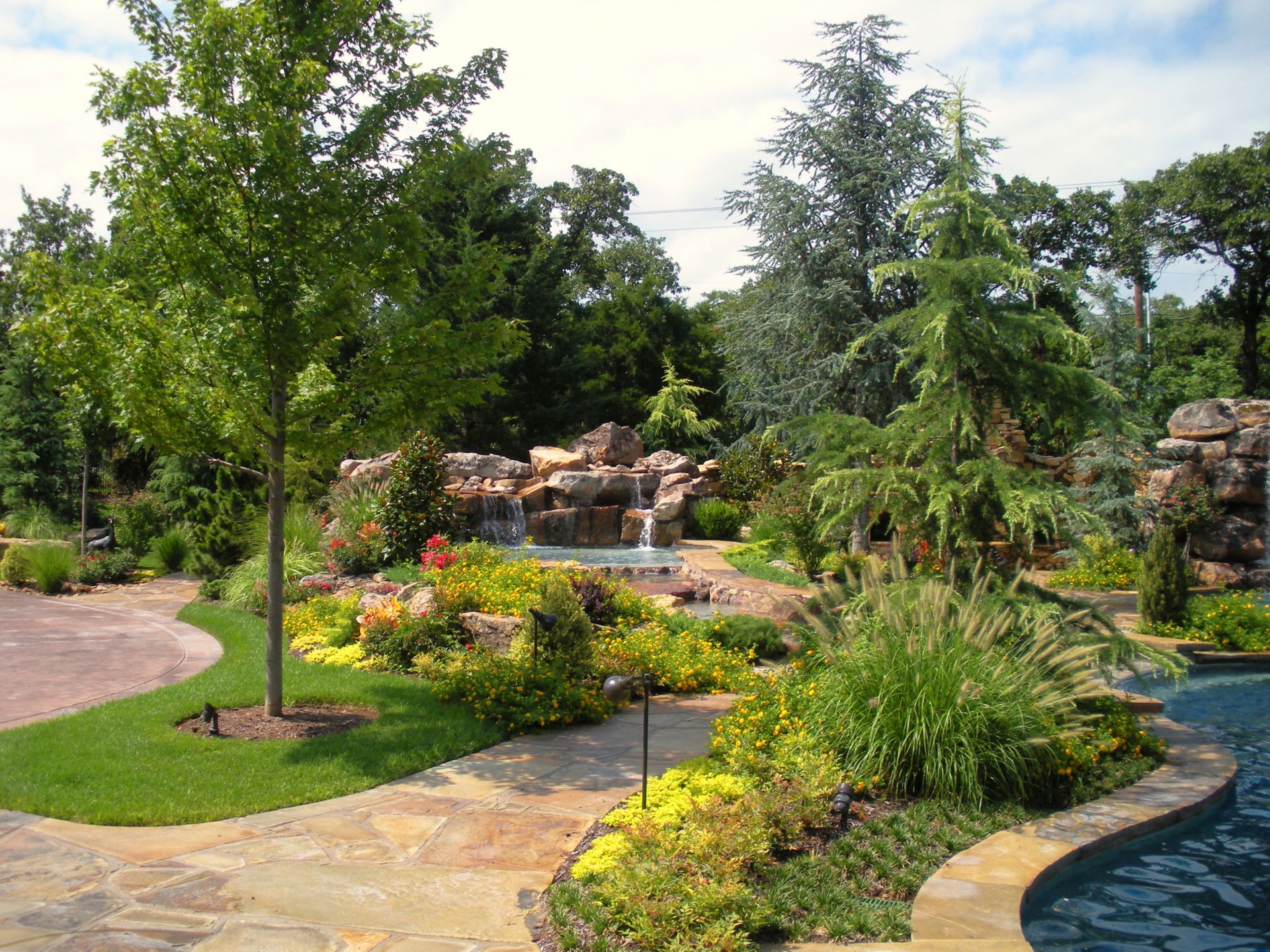 Landscaped garden with waterfall, trees, flowers, and a stone walkway surrounding a pool.