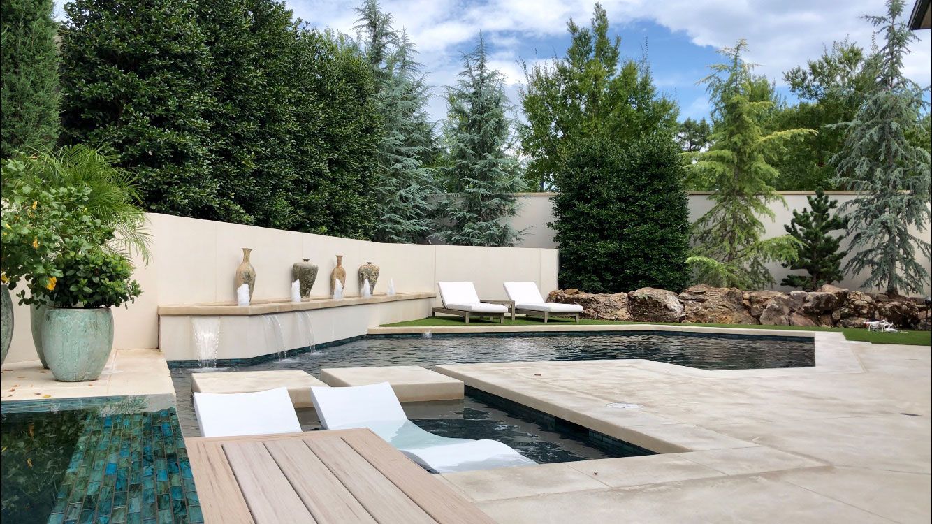 Pool area with lounge chairs, a shallow pool section, and greenery against a light-colored wall.