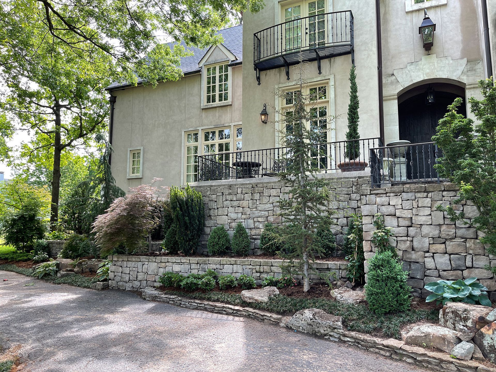 A stone-walled, multi-level garden with landscaping in front of a two-story home. A black balcony and door are visible.