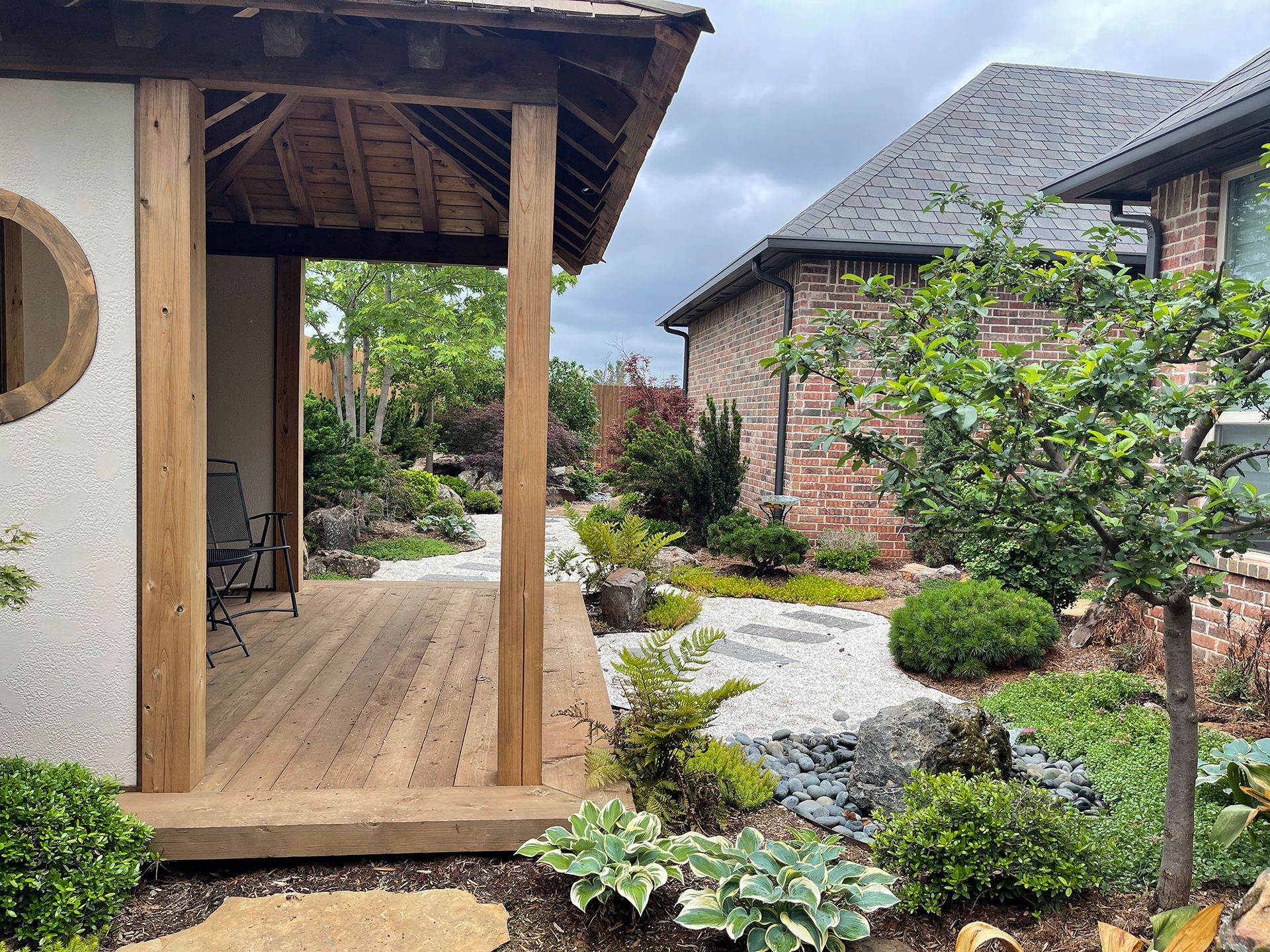 A wooden gazebo with a low roof overlooks a gravel path and a lush garden next to a brick house on a cloudy day.