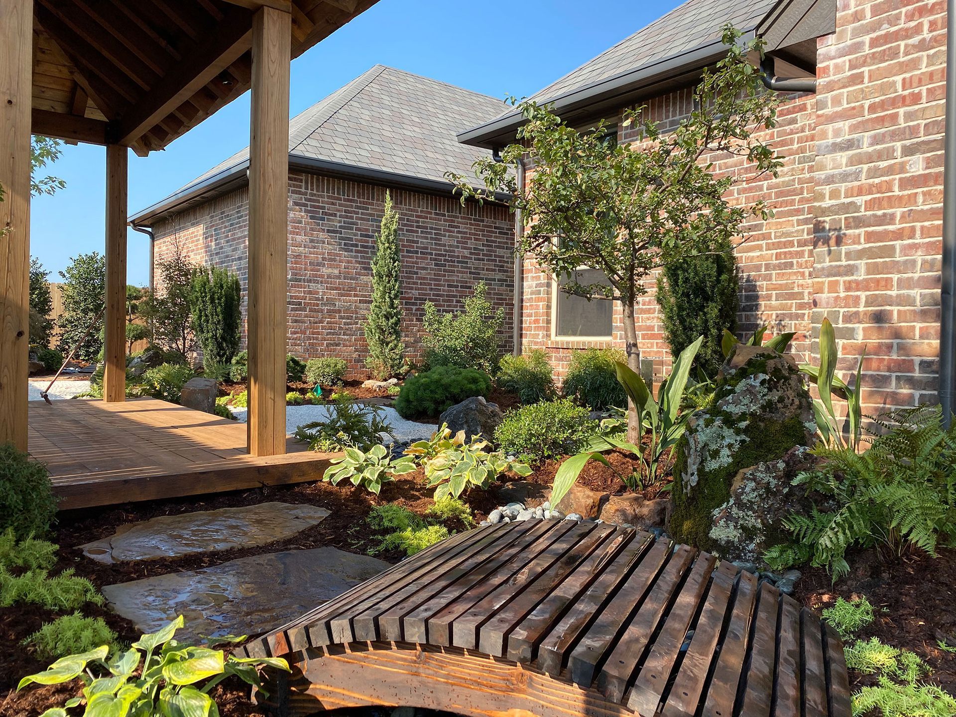 Wooden bridge and landscaped garden in front of a brick house with a covered porch.
