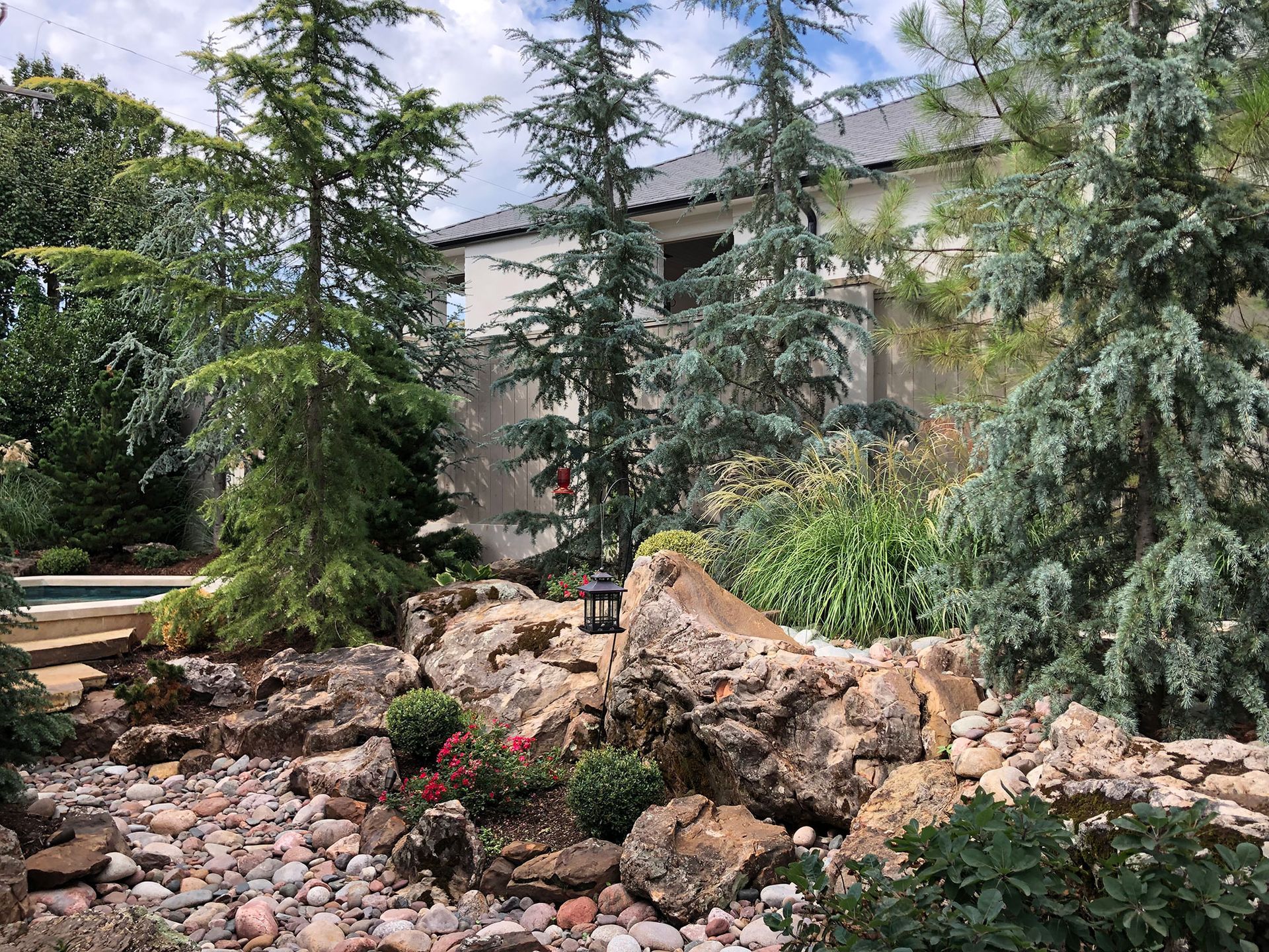 A rock garden with evergreens and various plants in front of a white building under a cloudy sky.