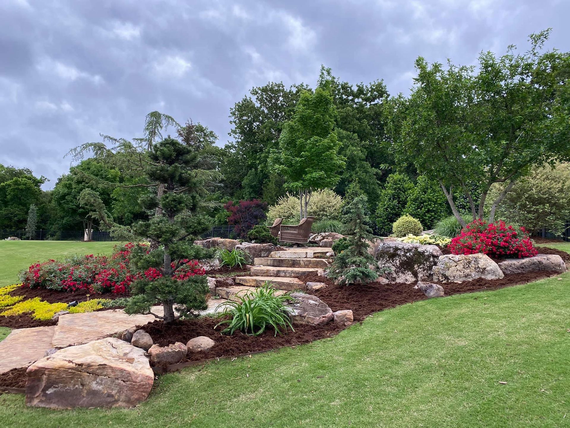 Stone steps lead up a garden bed filled with flowers, bordered by large rocks and lush greenery under an overcast sky.