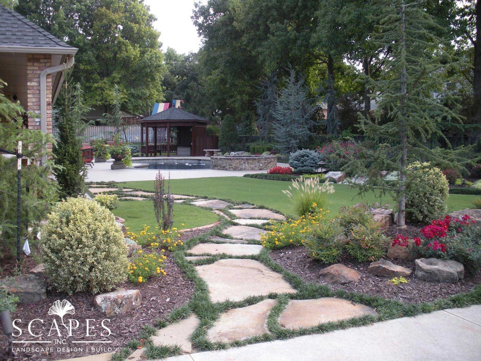 Stone path winds through a landscaped yard, leading to a gazebo and pool. Lush greenery, flowers, and trees surround the path.