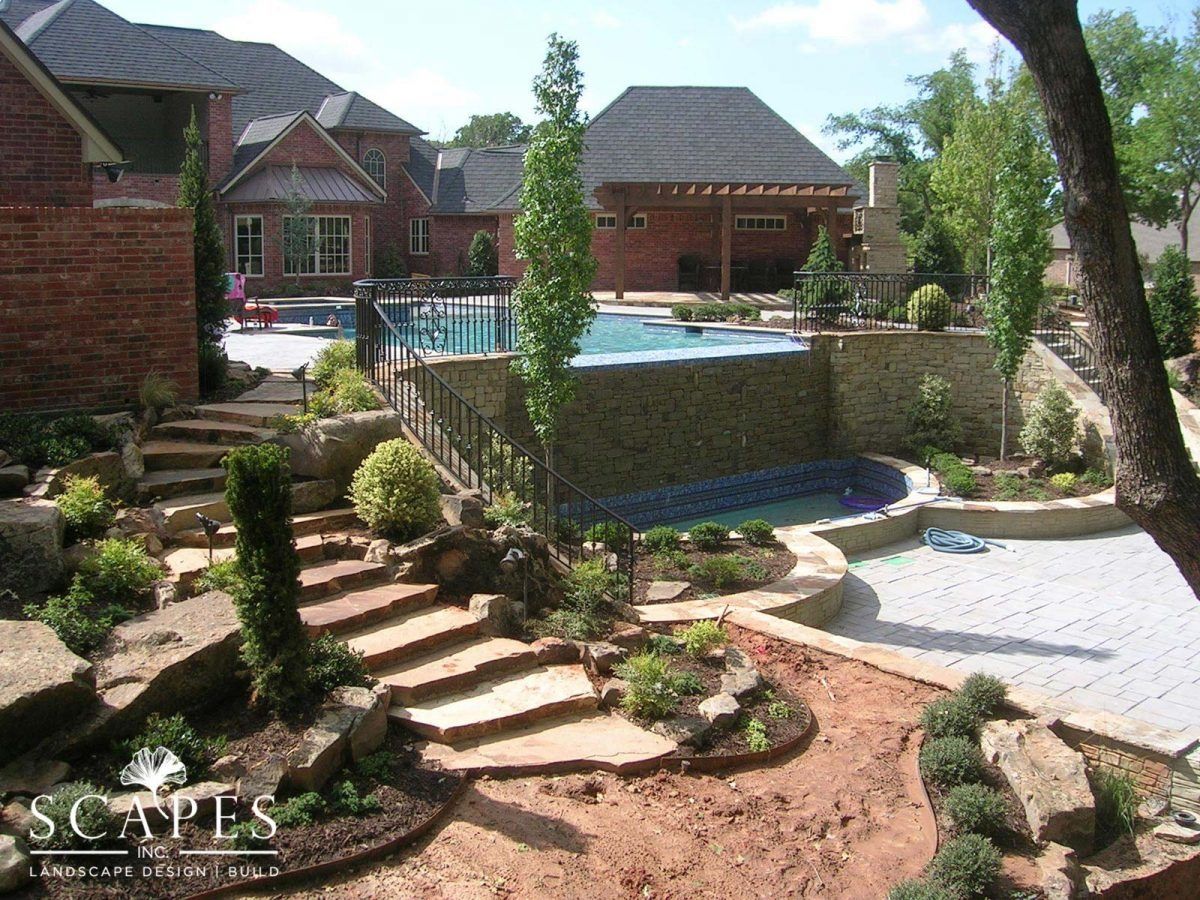Stone steps lead up to a pool and patio area surrounded by landscaping, with a large brick home in the background.