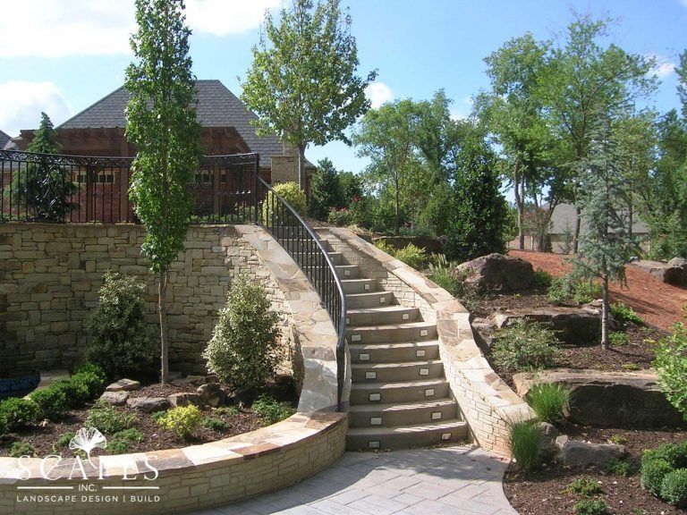 Stone steps with a curved handrail lead uphill through a landscaped garden. Trees and shrubbery flank the stairs and surrounding structures.