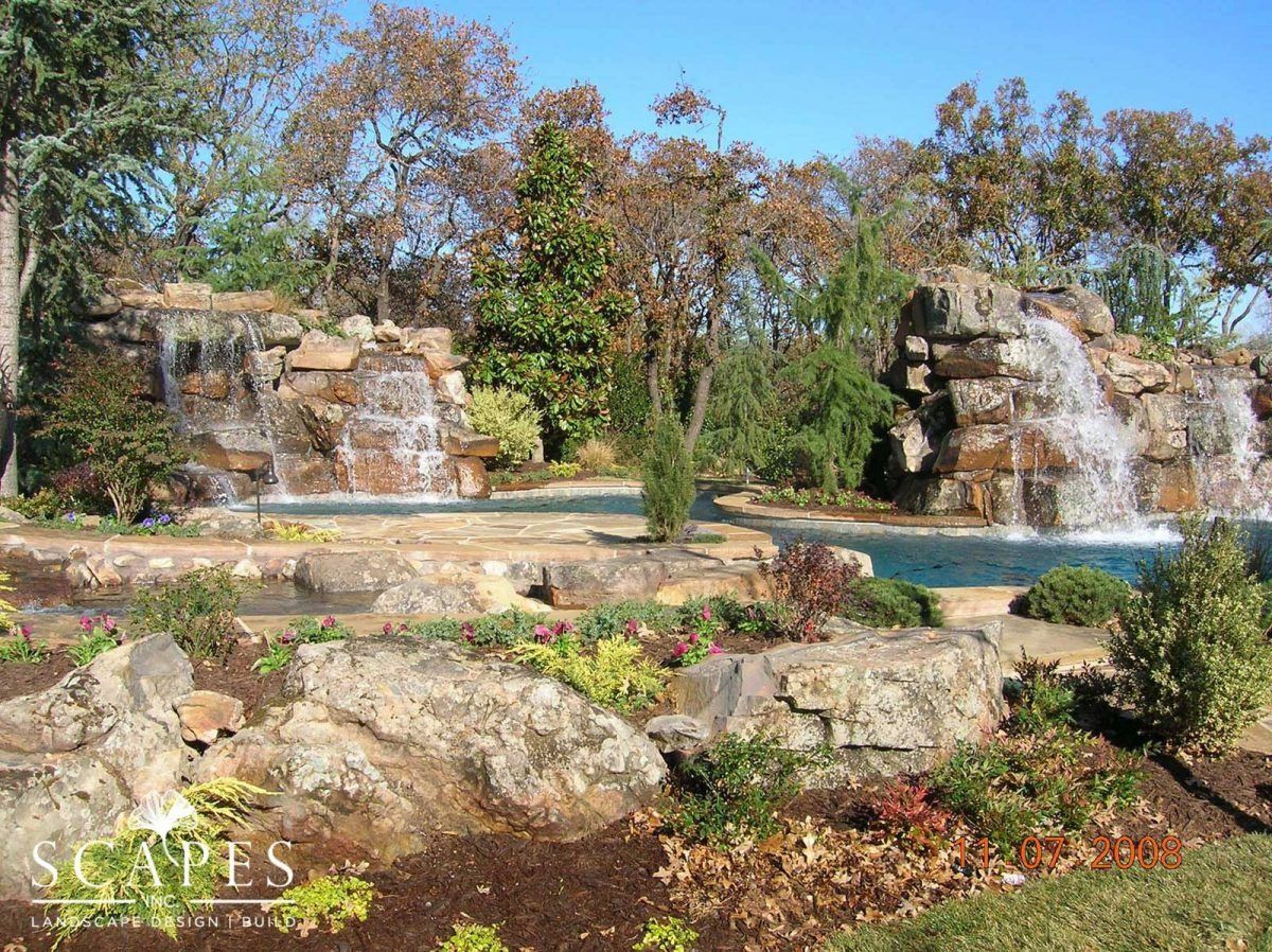 Two tiered waterfalls cascade into a pool surrounded by large rocks and landscaping. Blue sky overhead.
