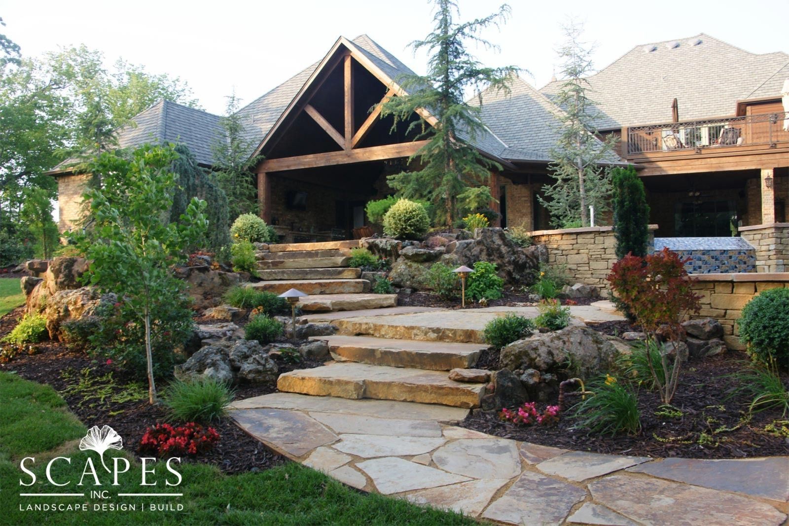 Stone steps and pathway lead to a covered outdoor kitchen and seating area, surrounded by landscaped plants and shrubs.