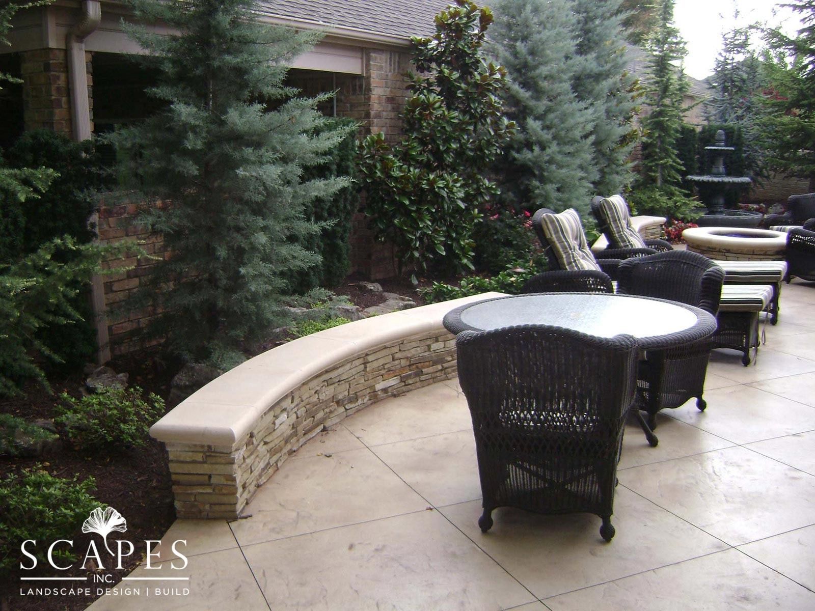 Outdoor patio with wicker furniture, a curved stone bench, and lush landscaping. A fountain is visible in the background.