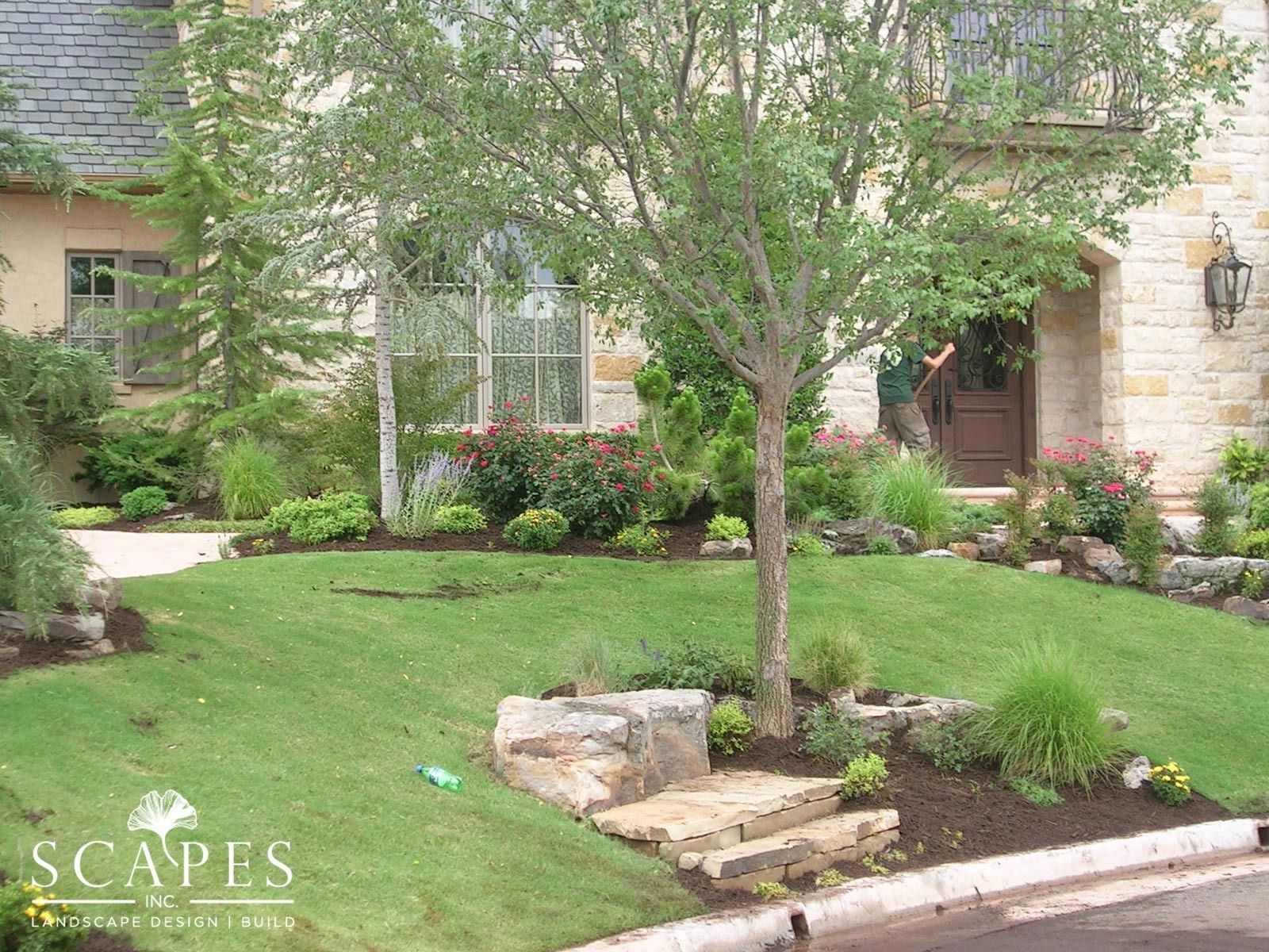 Lush green lawn with a tree in front of a stone house. Landscaping with flowerbeds and stone steps.