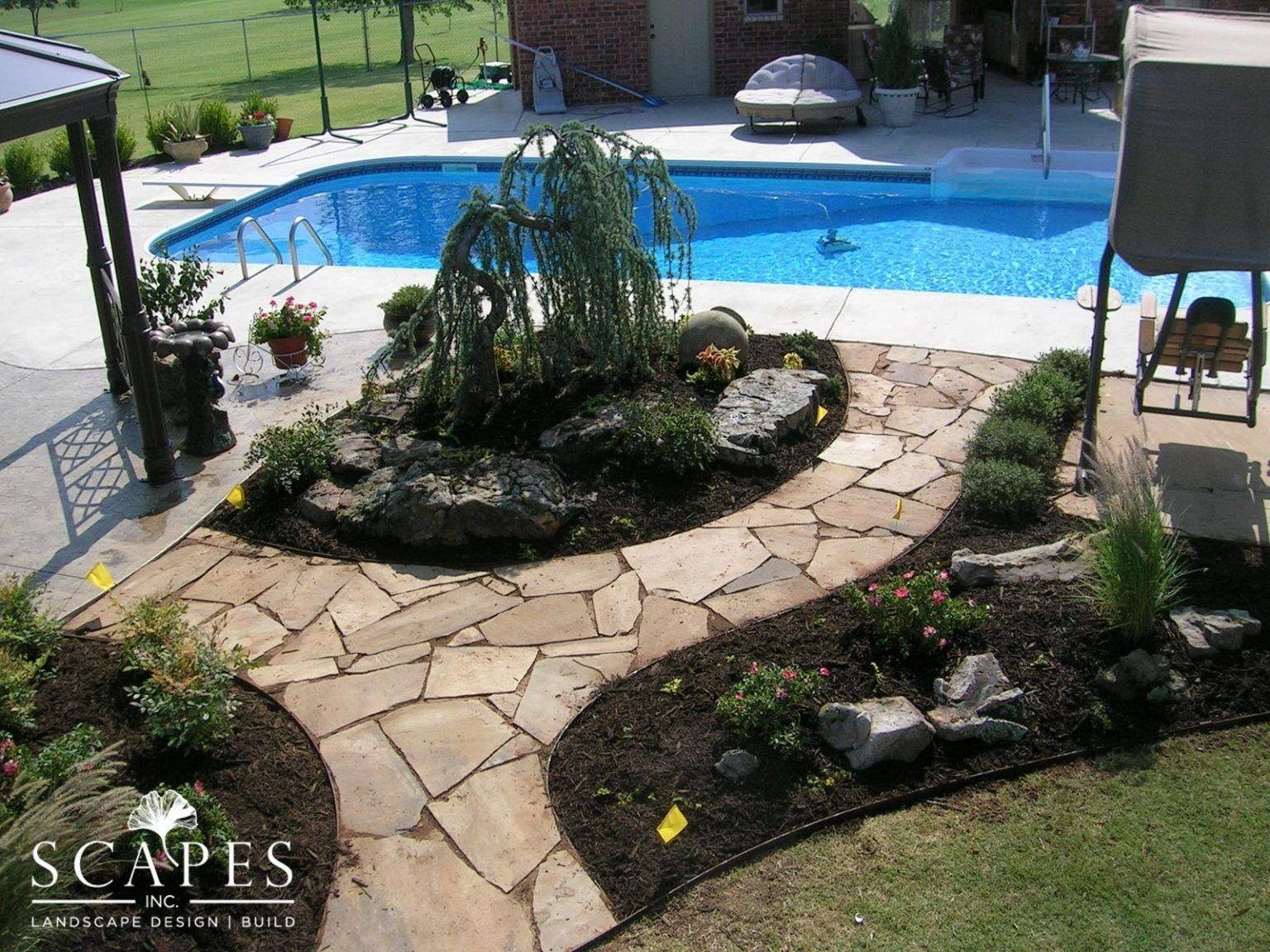 Flagstone pathway curves around a garden island with a weeping tree, leading to a swimming pool.