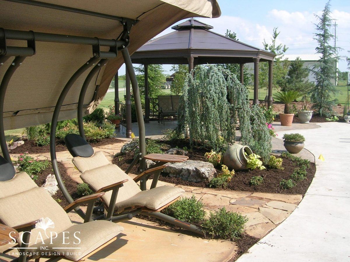 Outdoor scene with lounge chairs under a canopy, stone patio, garden with gazebo in the background.