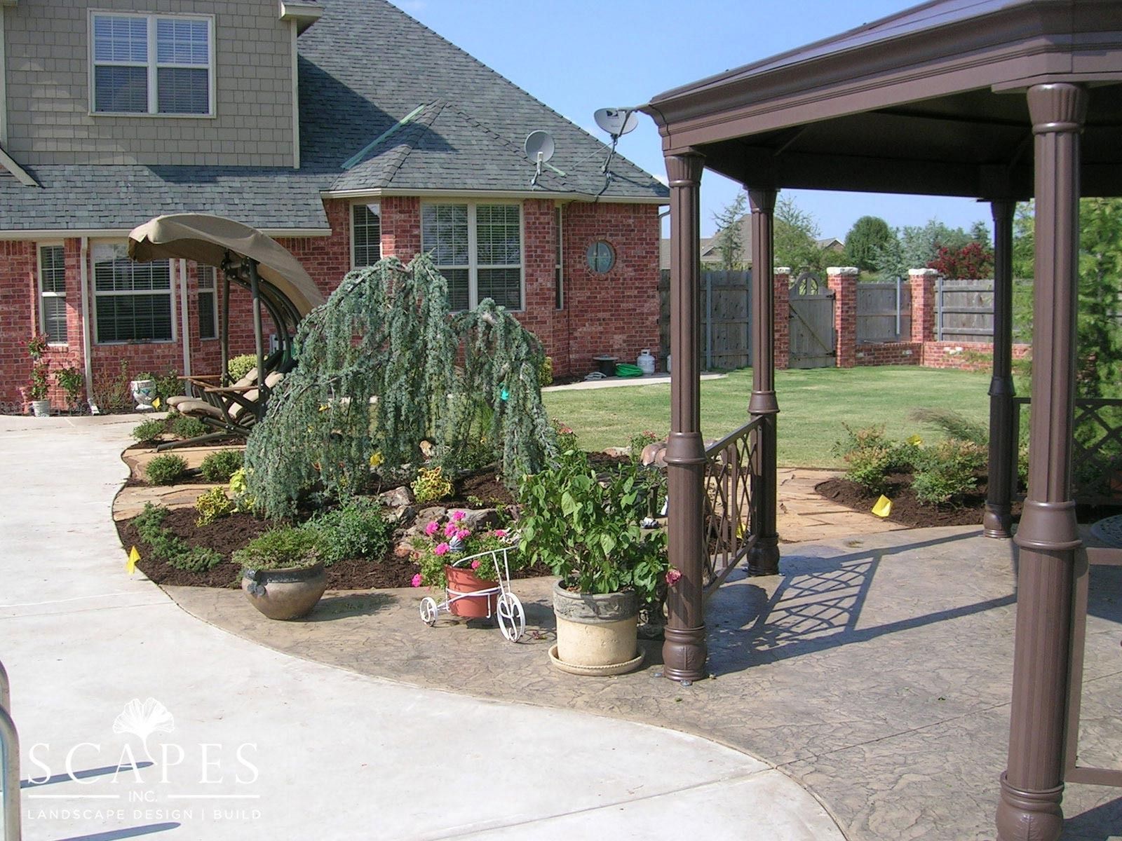 Landscaped backyard with a gazebo and weeping tree, curving pathway, and brick house in the background. Sunny day with a green lawn.