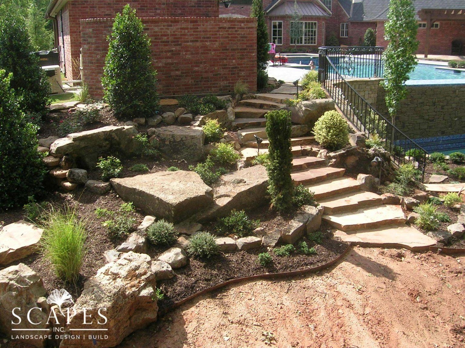 Stone steps and landscaping lead from a red brick building to a pool. Rocks and various plants are placed throughout the hillside.