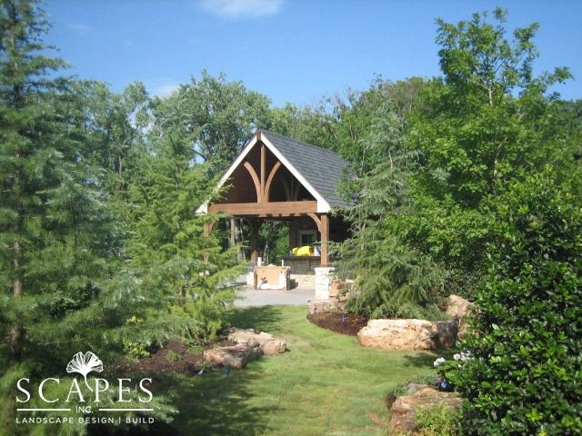 Wooden pavilion nestled among trees, with a path leading towards it. Lush greenery surrounds the structure.