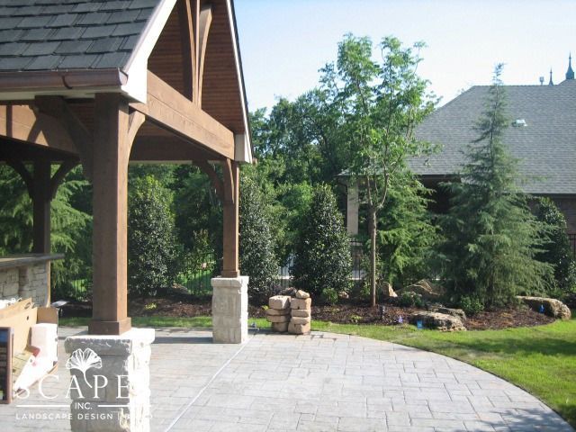 Patio with wooden pergola and a stone base, a paved area, with lush green trees in the background, and a house with a brown roof.