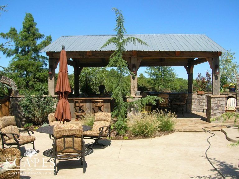 Outdoor patio with rustic wooden pergola, seating, and landscaping under a blue sky.
