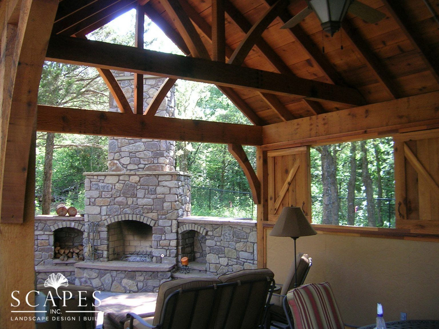 Wooden-framed outdoor living space with stone fireplace and comfortable seating, overlooking a forest.
