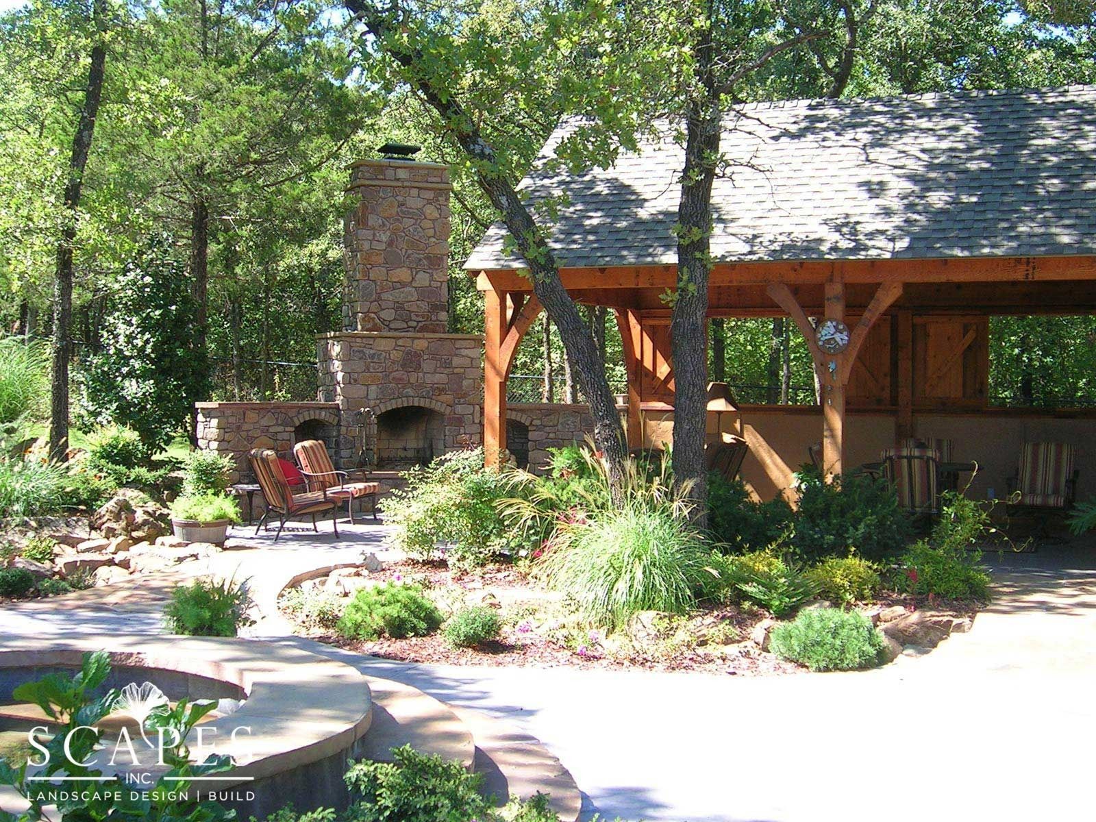 Outdoor living space with a stone fireplace, wooden pavilion, and lush landscaping.